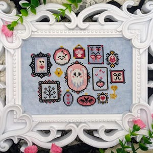 May include: A framed cross-stitch sampler featuring various small, framed images with pink, black, and white designs on a light blue background. The white frame has ornate details and is decorated with pink flowers and greenery.