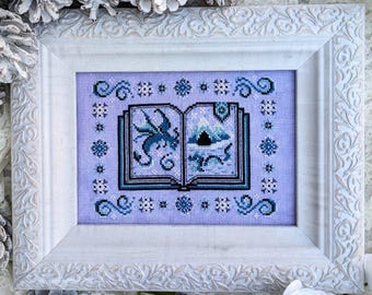 May include: A white frame with an embroidered image of an open book with a blue dragon inside. The book is on a purple background with snowflakes and swirls.