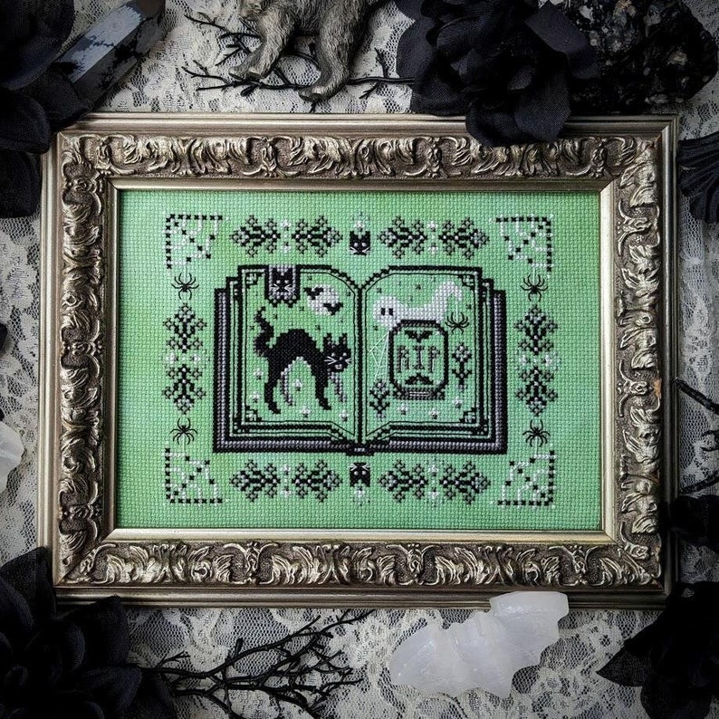 Book of Scaredy Cats cross stitch pattern. Features a spooked black cat coming across a cute spoopy ghost in the cemetery. Features a border of grey florals and spider webs.