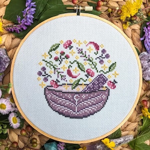 May include: A purple bowl filled with a colourful arrangement of flowers, herbs, and berries. The bowl is surrounded by a white background and a wooden frame.