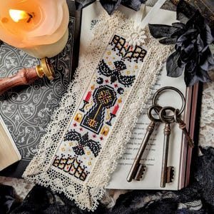 May include: A cross-stitched bookmark with a gothic design featuring a key, bats, and candles. The bookmark is white with black, red, and yellow accents, and is trimmed with cream lace. Three antique keys rest on a book.