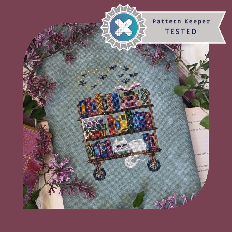 May include: A cross-stitch embroidery on mottled blue fabric featuring a three-tiered rolling cart filled with colorful books and a white cat resting on the bottom shelf. Above the cart, small black bats fly in a curved formation. Text reads Pattern Keeper TESTED.
