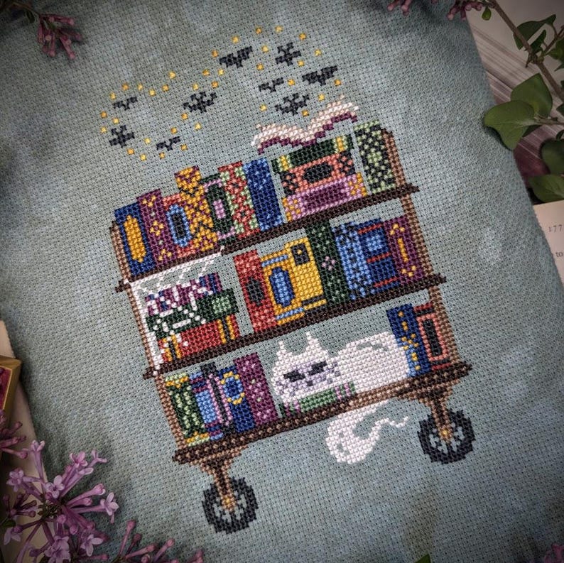 May include: A cross-stitch embroidery on light blue fabric featuring a three-tiered rolling cart filled with colorful books. A white cat sits on the bottom shelf, and small black bats and yellow stars are stitched above the top shelf.