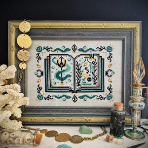 May include: Cross-stitch art of an open book with teal and black designs, framed in a gray and gold frame. The book features a trident and botanical elements. Decorative gold jewelry and glass bottles are in the foreground.