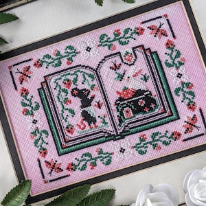 May include: A pink cross-stitch embroidery of an open book with a black mouse holding a strawberry. The book is surrounded by green vines and red strawberries. The background is a light pink with a black frame.