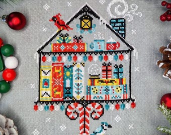 May include: A cross-stitch pattern of a house with a candy cane tree, presents, and a bluebird. The house is decorated with red, white, and blue colors. The pattern is on a gray background.