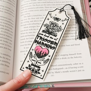 May include: A white bookmark with a black tassel, featuring the text "You had me at TRIGGER WARNING" and a pink heart with flames. The bookmark also includes illustrations of hands, flowers, and a butterfly.