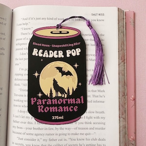 May include: Black bookmark shaped like a drinks can with a purple tassel. The bookmark features the text "Reader Pop" and "Paranormal Romance" with a full moon, wolf, and bat graphic. The can reads "Blood Moon - Shapeshifting Elixir" and "375ml".