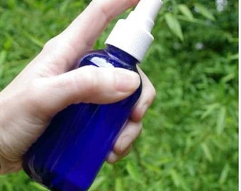 Puede incluir: Una botella de vidrio azul con un pulverizador blanco llena de aceite de magnesio. La botella está siendo sostenida por una mano frente a un fondo verde frondoso.
