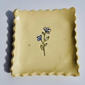 Puede incluir: Un plato cuadrado de cerámica con borde festoneado. El plato es de color amarillo pálido y presenta un diseño de flor azul pintado a mano. La flor tiene dos flores y un tallo verde. El plato probablemente se utiliza para servir artículos pequeños.