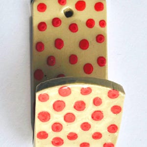 May include: A ceramic wall hook with a cream-colored base and red polka dots. The hook has a rectangular back with a hole for hanging and a curved front section. The edges are painted dark green.