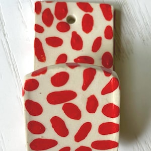 May include: A ceramic wall pocket with a cream-colored base and a pattern of red, irregular shapes. The pocket has a rectangular top section with a hole and a curved bottom section.