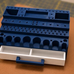 May include: A dark blue plastic organizer stand featuring multiple rows of circular and rectangular slots for tool storage. The unit includes a single white sliding drawer at the base with a dark blue handle, providing additional storage space.
