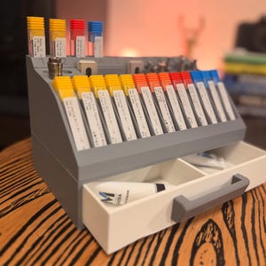 May include: A gray 3D-printed storage organizer with a white pull-out drawer containing small tubes. The top section features angled slots holding multiple clear plastic cases with yellow, orange, and blue caps, each labeled with text.
