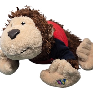 Puede incluir: Un mono de peluche marrón y beige que lleva una camisa roja y azul. El juguete tiene una cabeza peluda marrón y una cara beige con una nariz negra. El juguete tiene una etiqueta de Webkinz en su pie.