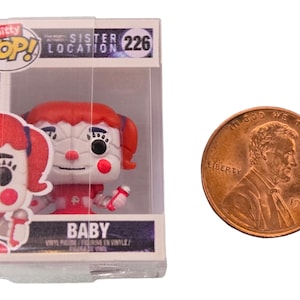 Puede incluir: Una figura de vinilo Funko Pop! de Baby del juego Sister Location. La figura está en una caja de plástico transparente con un diseño rojo, blanco y azul. La caja tiene las palabras "Sister Location" y "226" en ella. La figura es aproximadamente del tamaño de una moneda de un centavo de euro.