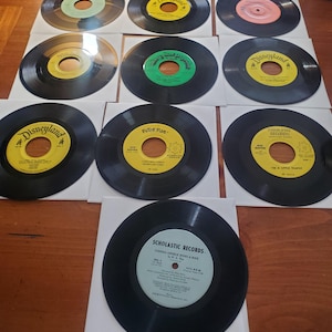 May include: A collection of vintage 45 RPM vinyl records. The records have colorful labels with text, including "Disneyland," "Peter Pan," and "Scholastic Records." The records are in white paper sleeves, arranged on a wooden surface.