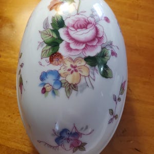 May include: A white, egg-shaped decorative porcelain piece adorned with a large pink rose and other colorful floral designs. Green leaves and smaller flowers in blue, yellow, and orange accent the main floral arrangement. The egg has a gold trim.