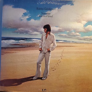 May include: Album cover for Bobby Goldsboro's "Summer (the first time)". The artwork depicts a man in a light-colored suit standing on a beach with footprints in the sand. The sky is blue with white clouds, and the ocean is visible in the background.