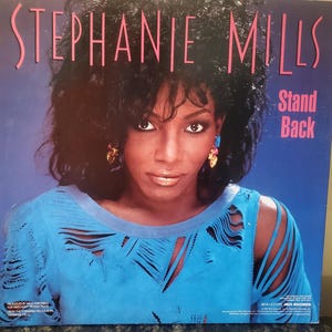 May include: Album cover for Stephanie Mills' album, featuring the text "STEPHANIE MILLS" in pink. The album title "Stand Back" is in white. Mills is wearing a blue top with cut-out details and gold earrings. Her dark hair frames her face.