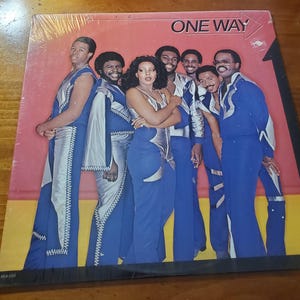 May include: Album cover featuring the group One Way. The group is composed of seven people, all wearing blue and silver outfits. The album cover has the words "ONE WAY" in white letters on a red background.