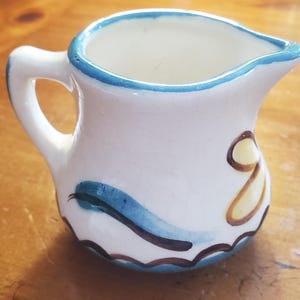 May include: A small, white ceramic creamer with a handle and spout. The rim is trimmed with blue, and the body is decorated with blue and brown wave-like patterns and a yellow floral design. The creamer is approximately 3 inches tall.