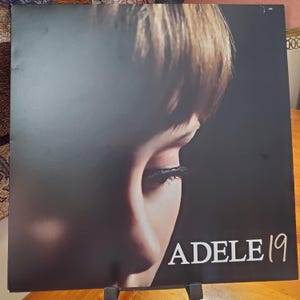 May include: Album cover for Adele's debut album, "19." The cover features a close-up of Adele's face, with her eye and part of her hair visible. The album title and artist name are in white text.