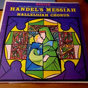 May include: A vintage record album cover featuring a stained glass style illustration of two figures embracing. The album is titled "Handel's Messiah" with the text "featuring the famous Hallelujah Chorus" below. The background is a vibrant purple.