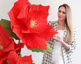Könnte beinhalten: Eine Frau steht vor einer großen, roten Papier-Weihnachtsstern-Blume mit grünen Blättern. Der Text "DIY Giant Poinsettia with Lights and Curvy Stem" befindet sich auf einem roten Hintergrund.
