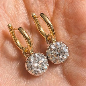 Op de afbeelding: Een paar gouden oorringen met grote, ronde, sprankelende diamanten hangers. De diamanten zijn gezet in een zilverkleurige bezel, tegen een huidkleurige achtergrond.