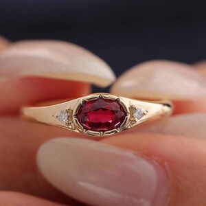 Könnte beinhalten: Ein Goldring mit einem zentralen, oval geschliffenen, tiefroten Rubin in einer Zargenfassung, flankiert von zwei kleinen, rund geschliffenen, klaren weißen Diamanten auf beiden Seiten der Ringschiene für ein symmetrisches Design.
