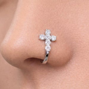 May include: A silver nose ring featuring a cross design, adorned with sparkling clear gemstones. The cross is composed of five gemstones, with three additional gems below. The jewelry is shown on a person's nose.