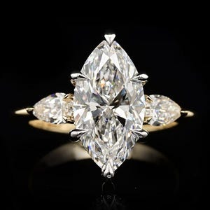 May include: A gold ring featuring a large marquise-cut center stone held by silver-toned prongs, flanked by two smaller pear-cut stones on each side. The band is a polished yellow gold, and the setting provides a clear view of the faceted stones.