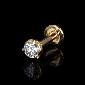 May include: A gold flat back labret stud featuring a single round brilliant cut clear gemstone held in a four-prong setting. The post is smooth and cylindrical, ending in a circular, textured screw-on backing piece with a metallic finish.