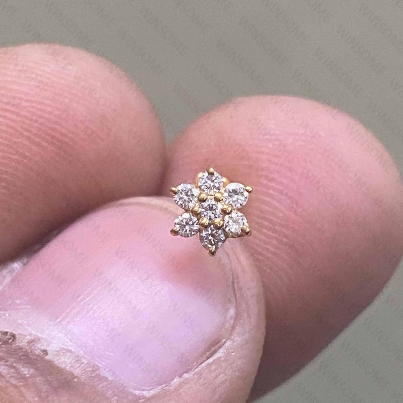 May include: A small, gold-colored flower-shaped stud earring. The earring features seven clear, round-cut gemstones set in a floral pattern. The earring is held between two fingers.