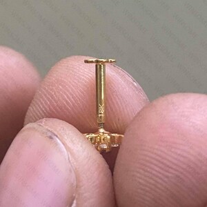 May include: A gold stud earring with a flat back post featuring the engraving 18K on the stem. The earring has a decorative front component with a clear stone setting, and the post is designed for secure insertion through a piercing.