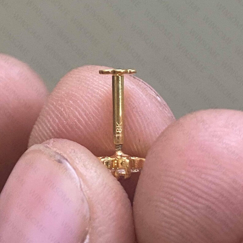 May include: A gold stud earring with a flat back post featuring the engraving 18K on the stem. The earring has a decorative front component with a clear stone setting, and the post is designed for secure insertion through a piercing.