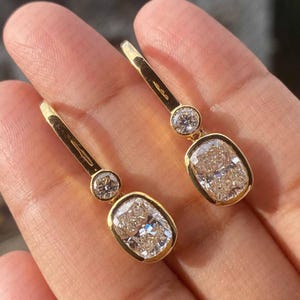 May include: A pair of gold drop earrings featuring a small round clear stone set above a larger rectangular cushion-cut stone with rounded corners. The earrings have a polished gold finish and a secure lever-back closure mechanism for attachment.