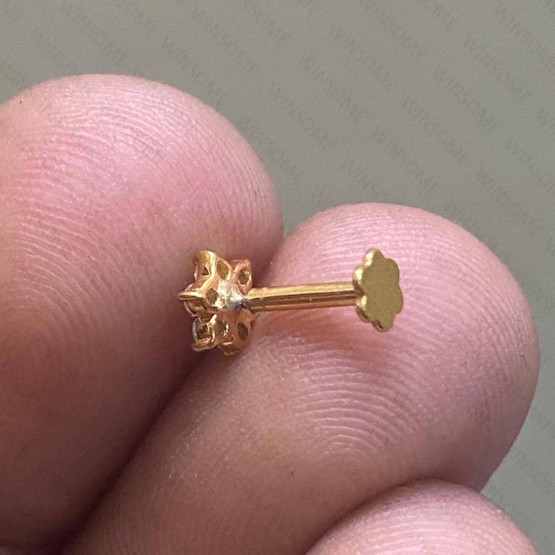 May include: A gold-colored stud earring with a flower-shaped design on one end and a flat, petal-shaped backing. The earring is held between fingers, showcasing its intricate details and polished finish. The jewelry piece is designed for ear piercing.