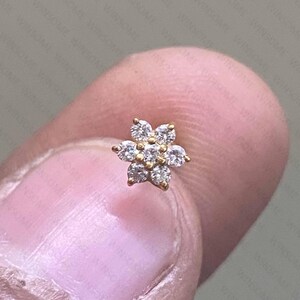 May include: A gold stud earring featuring a flower-shaped cluster of seven small, clear, round-cut gemstones. The central stone is surrounded by six petals, each set with a single sparkling stone, creating a delicate floral arrangement.