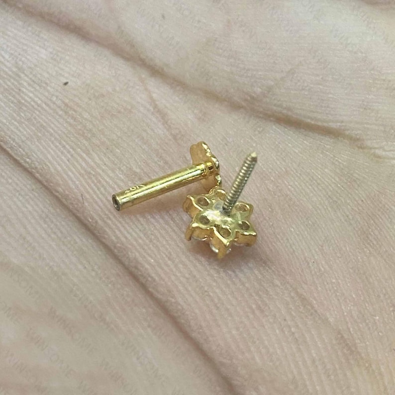 May include: A gold stud earring featuring a flower-shaped head with six clear stones arranged in a circular pattern. The earring has a threaded post and a flat, circular back piece, with the text 18K engraved on the post.