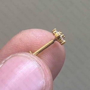 May include: A gold-colored, flower-shaped stud earring with a small post. The earring is held between fingers, showcasing its delicate design and intricate details. The stud's floral design is accented with small, sparkling stones.