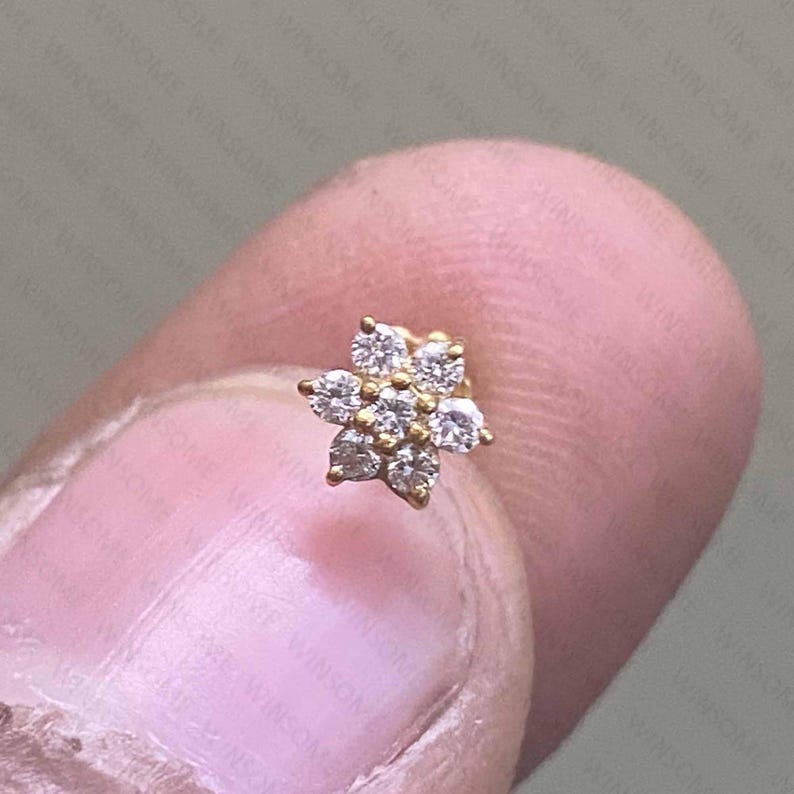 May include: A gold stud earring featuring a flower-shaped cluster of seven small, clear, round-cut stones. The central stone is surrounded by six petals, each formed by a single stone, creating a symmetrical floral design.