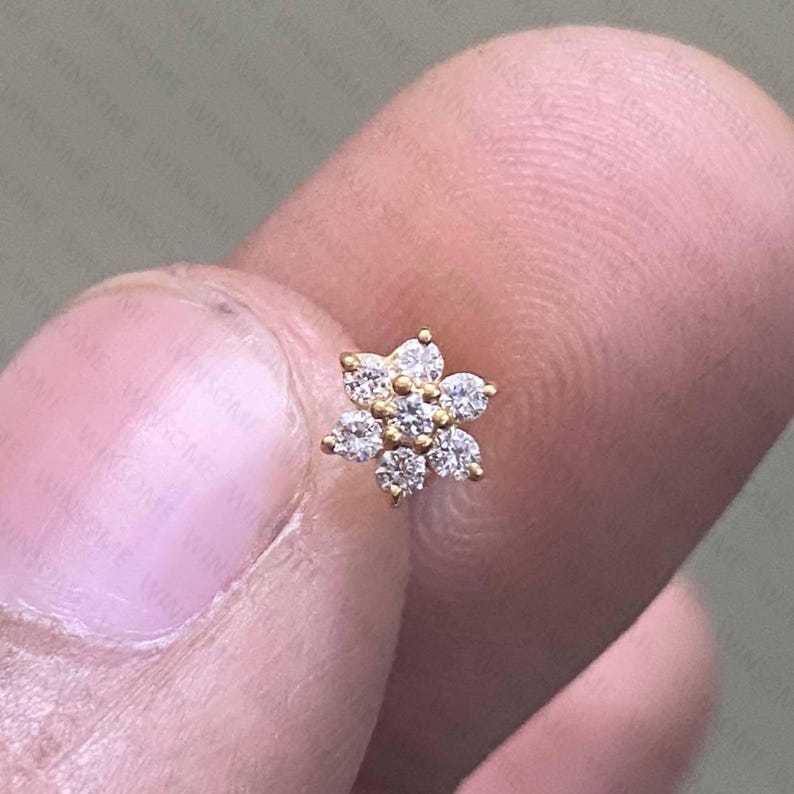 May include: A small, gold-colored flower-shaped stud earring. The earring features six clear, round-cut gemstones arranged around a central gemstone. The earring is held between two fingers.