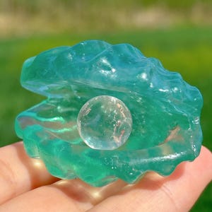May include: A green glass seashell with a clear glass pearl inside. The seashell is open and the pearl is visible.