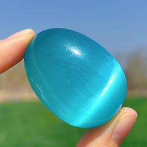 Op de afbeelding: Een gladde, gepolijste, lichtblauwe edelsteen in de vorm van een ei. De edelsteen heeft een subtiel schimmeren en wordt in een hand gehouden.