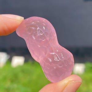 May include: A pink, translucent gemstone carved in the shape of a peanut. The stone is held between two fingers, with a blurred green background. The gemstone has a textured surface and a soft, rosy hue.