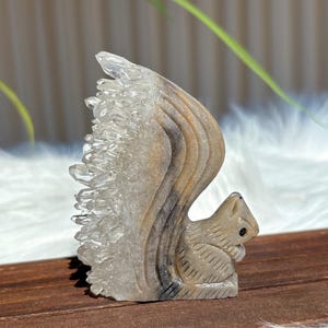 Puede incluir: Una figurita de ardilla tallada en cristal con una cola tupida de cristales de cuarzo transparentes. La ardilla está sentada sobre una superficie de madera.