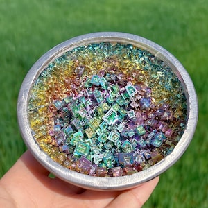 Può includere: Una ciotola d'argento piena di cristalli iridescenti con colori arcobaleno. I cristalli sono piccoli e di forma quadrata, creando un effetto scintillante.