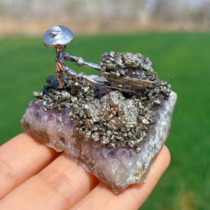 以下が含まれることがあります： アメジストの結晶のベッドの上に、ピライトの結晶でいっぱいの手押し車を押している銀の置物です。